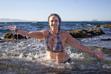 Young brunette woman in a bikini bathing on a rocky beach with water splashing on her.