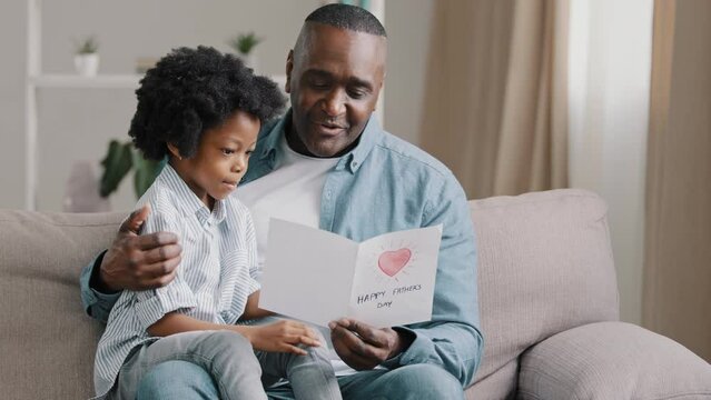 Mature african american man with cute kid girl hugging sitting in room on sofa father reads greeting card loving daughter made gift with own hands for birthday or father's day dad happy cuddling baby