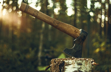 An axe with a wooden handle is stuck in a wooden log. Logging on the background of sunset. The...