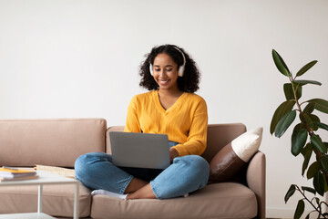 Young happy black woman in headphones using laptop pc, working online, sitting on couch at home, copy space