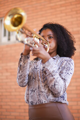 Front low angle view of a young female street musician playing the trumpet with a brick wall...