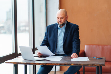Bald adult businessman beard working doing paperwork, in suit jacket, sitting wooden desk laptop, signing contract, deal, smartphone, light window. Light brown sofa back office. Not pleased agreement