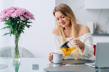 Blond woman kitchen pouring drinking yellow orange diet herb tea teapot. Wearing knitted sweater sitting glass table, bouquet flowers vase. Preparing working day. Blurred background kitchen-ware