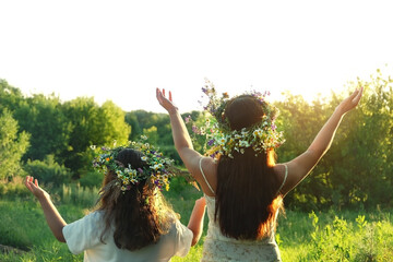 Two girls in flower wreaths on meadow, sunny green natural background. Floral crown, symbol of summer solstice. Slavic ceremony on Midsummer, wiccan Litha sabbat. pagan holiday Ivan Kupala - obrazy, fototapety, plakaty