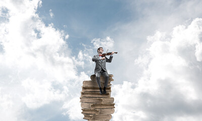 Handsome violinist in blue day sky on pile of books play his melody