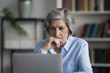 Pensive focused elderly mature 50s business woman looking at computer screen, reading email,...