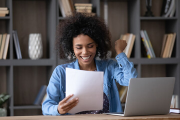 Naklejka na ściany i meble Joyful laughing young African American businesswoman feeling excited reading paper document, celebrating professional success, getting dream job offer or win news, sitting at table at home office.