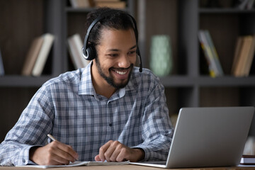 Happy young African American man in wireless headphones with microphone looking at laptop screen, enjoying improving knowledge watching educational lecture online, e-learning preparing for exam.