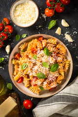 Italian pasta alla arrabiata with basil and parmesan cheese on dark table with ingredients. Fusilli pasta with tomato sauce arrabbiata. Top view, vertical.