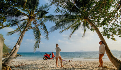 Scooter road trip. Lovely couple on red motorbike in white clothes on sand beach. Wedding just married people walking near tropical palm tree, sea. Honeymoon by ocean. Asia Thailand. Motorcycle rent.