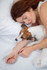 Caucasian red-haired woman sleeps in an embrace with a jack russell terrier dog on a white sheet.