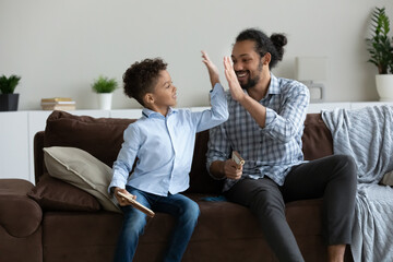 Happy little cute African American kid boy giving high five to caring millennial father, playing entertaining war game with wooden guns, enjoying domestic hobby activity pastime, having fun indoors.