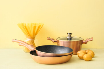 Copper frying pans, cooking pot, potatoes and jar with raw pasta on yellow background