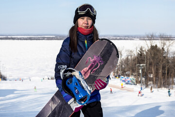A young beautiful woman in a ski mask stands on the top of a ski mountain with a snowboard in her...