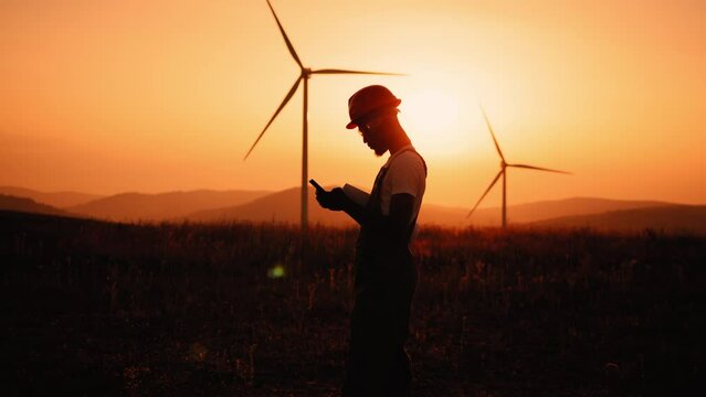 Silhouette African american industrial worker having mobile conversation while standing on farm with wind turbines. Engineer in helmet and uniform working on eco station. Man with wind turbines.