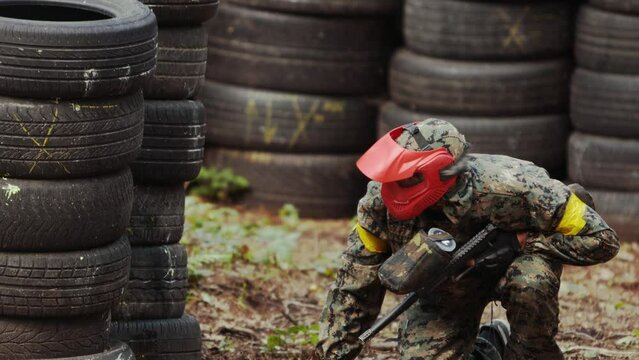 Portrait of american paintball player in mask who is aiming in opponents at war arena. Paintball player in a mask who is aiming at opponents. Man hides from the enemy behind tires