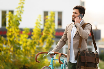 Young bearded businessman with bicycle talking by mobile phone in park