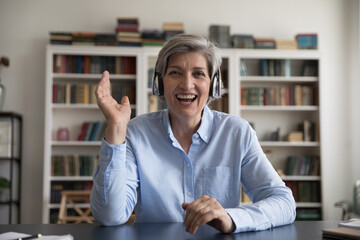 Web camera head shot view joyful attractive middle aged woman in wireless headphones waving hand,...