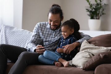 Cheerful affectionate 30s African American father showing funny videos online on cellphone to laughing adorable small kid daughter, enjoying playing games, posing for selfie photos, tech addiction.
