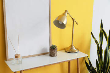 Table with golden lamp, frame, flowerpot and reed diffuser near yellow wall