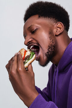 An African American man in a purple hoodie with short curly hair and beard open his mouth widely to bite a burger