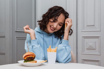 A horizontal photo of a pretty curly Asian girl wearing a blue hoodie sitting at the table looking...