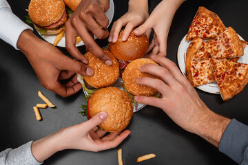 People’s hands reach out to the plate with four burgers, plates with burgers and pizza around them, french fries lie on the black table