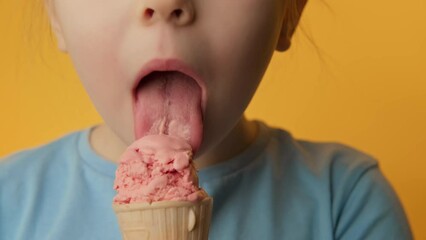 Happy toothless girl eating strawberry ice cream on yellow background. Child with ice cream looking at camera. Close-up in 4K, UHD