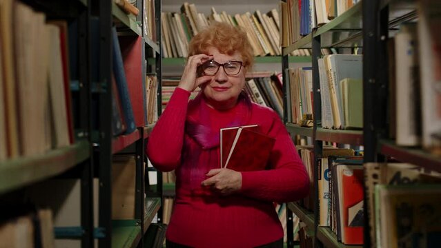 Smart elderly teacher in glasses holding books in her hands in the library