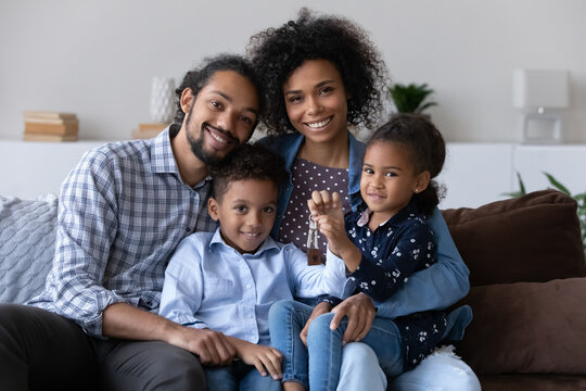 Portrait Of Happy Bonding African American Family Showing Keys To Camera, Sitting On Couch, Joyful Adorable Small Children And Caring Young Multiracial Couple Parents Celebrating Moving Into Own Home.