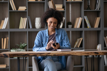 Happy beautiful young African American woman using cellphone, enjoying communicating in social networks, web surfing information, shopping or playing games, sitting at table in modern workplace.