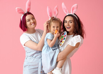 Young lesbian couple with little daughter, Easter egg and paints on color background