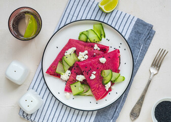 Beetroot pancakes folded into a triangle with cream cheese and fresh cucumber slices on a white plate on a light concrete background. Pancake recipes, international pancake day