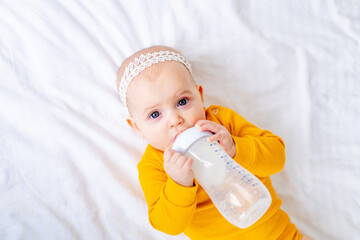 a baby sucks a bottle of milk holding in his hands on a white cotton bed in a yellow bodysuit, baby...