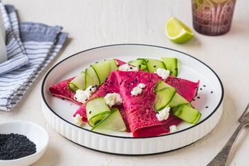Beetroot pancakes folded into a triangle with cream cheese and fresh cucumber slices on a white plate on a light concrete background. Pancake recipes, international pancake day