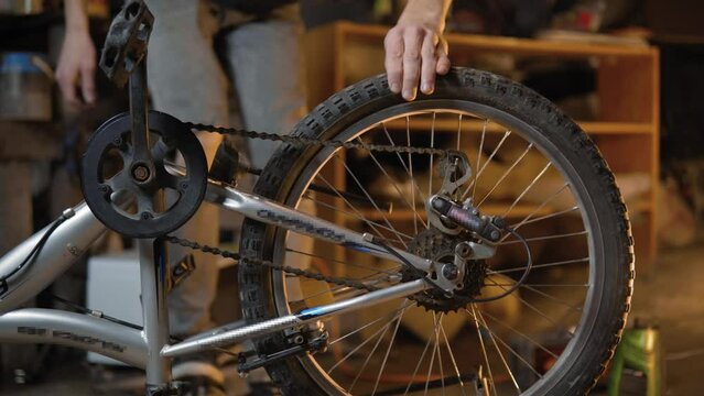 A slender guy repairs a broken bike in the garage. A man works in a room with old bicycle equipment. Repair of equipment with your own hands. The guy lubricates the chain and wheels with oil