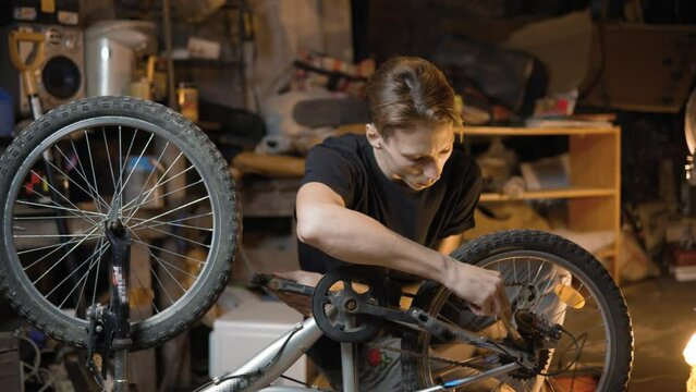 A slender guy repairs a broken bike in the garage. A man works in a room with old bicycle equipment. Repair of equipment with your own hands. The guy lubricates the chain and wheels with oil