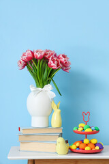 Vase with tulips, books, decor and Easter eggs on table near blue wall