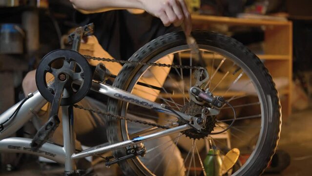A slender guy repairs a broken bike in the garage. A man works in a room with old bicycle equipment. Repair of equipment with your own hands. The guy lubricates the chain and wheels with oil
