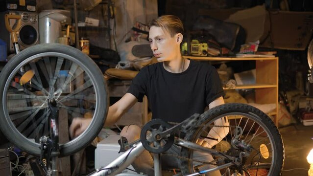 A slender guy repairs a broken bike in the garage. A man works in a room with old bicycle equipment. Repair of equipment with your own hands. The guy lubricates the chain and wheels with oil