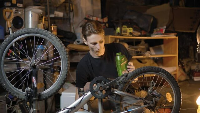 A slender guy repairs a broken bike in the garage. A man works in a room with old bicycle equipment. Repair of equipment with your own hands. The guy lubricates the chain and wheels with oil