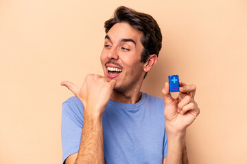 Young caucasian man holding a batterie isolated on beige background points with thumb finger away, laughing and carefree.