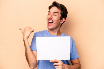 Young caucasian man holding a placard isolated on beige background points with thumb finger away, laughing and carefree.