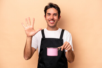 Young caucasian man holding a saucepan isolated on beige background smiling cheerful showing number five with fingers.