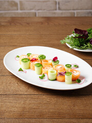 Appetizer of lightly salted salmon with tender cheese and cucumber on a white plate on a wood background. Beautiful appetizer for a festive party. Rolls of red fish and cucumber. Copy space. Close up.