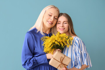 Young woman with her mother, gift and flowers on blue background