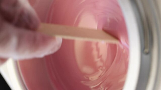 Process of depilatory wax heated and melted in a special device. Beauty master mixes the heating wax with a wooden spatula before the depilation waxing procedure. Vertical video