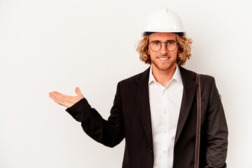 Young architect caucasian man with helmet isolated on white background showing a copy space on a palm and holding another hand on waist.