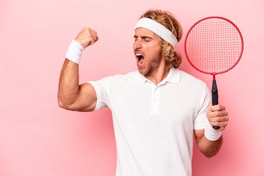 Young caucasian man playing badminton isolated on pink background raising fist after a victory, winner concept.