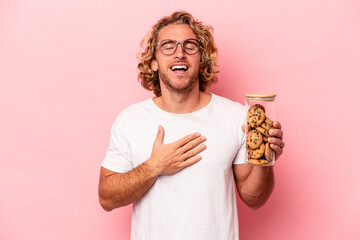 Young caucasian man holding cookies jar isolated on pink background laughs out loudly keeping hand...